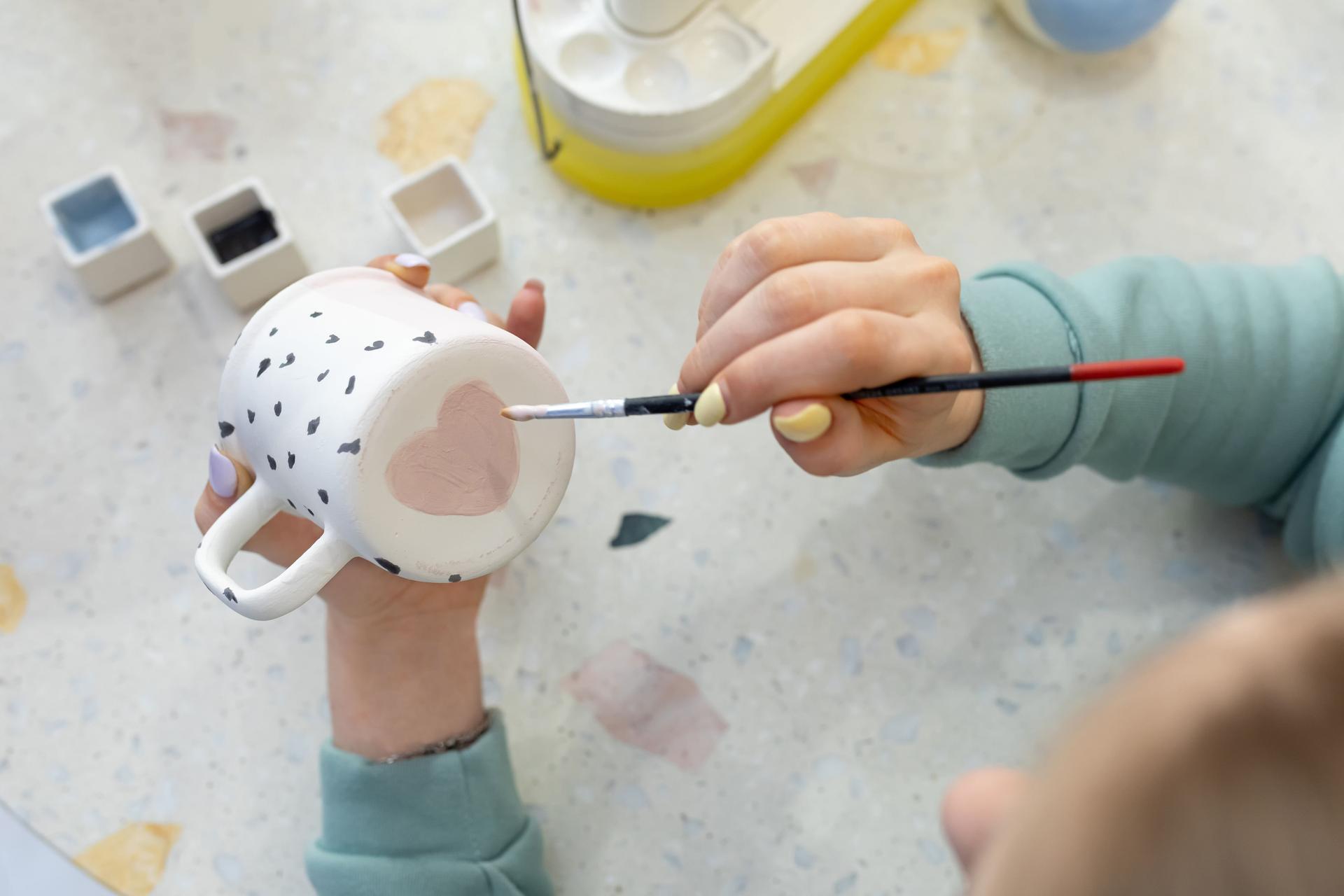 Women's hands paint pink heart on back of ceramic cup with brush. Training in profession of ceramicist, improvement of skills. Ceramic art concept.